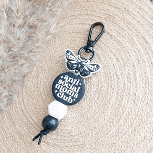 Op de afbeelding: Zwart-witte sleutelhanger met een ronde hanger met de tekst "anti-social moms club" en een zwarte vlinderhanger. De sleutelhanger heeft zwarte en witte kralen.