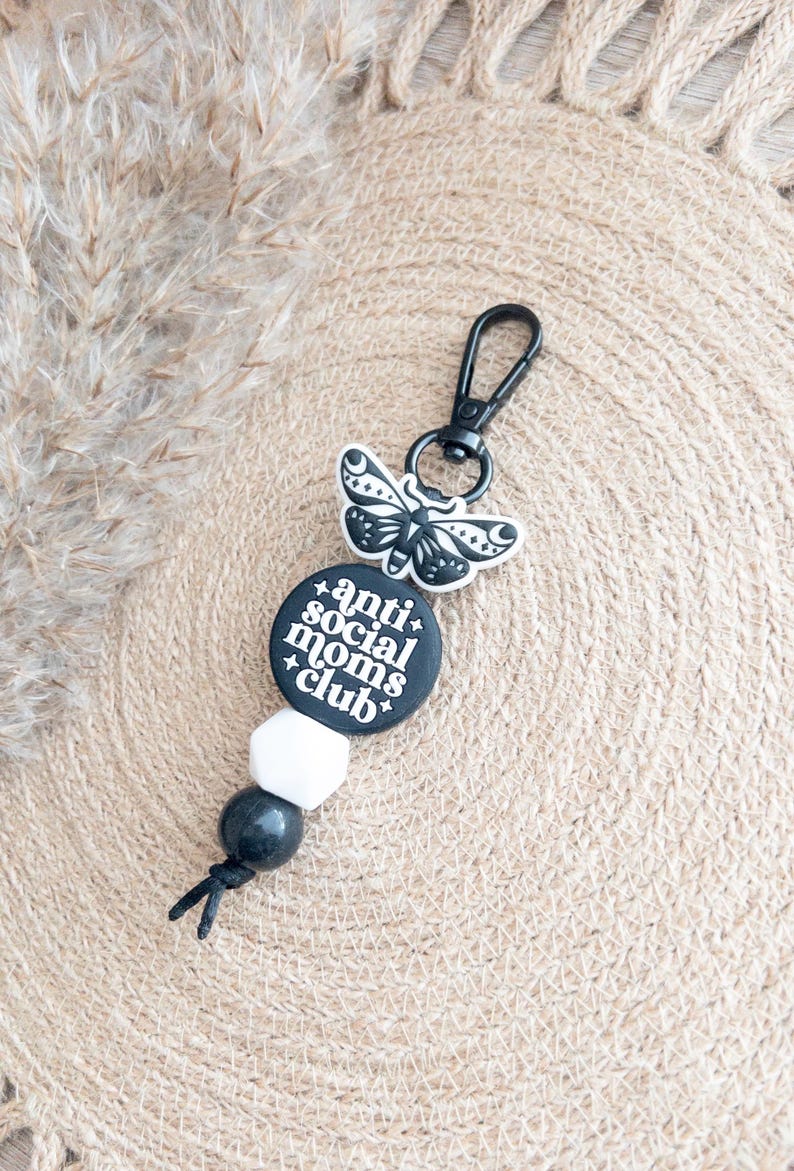 Op de afbeelding: Zwart-witte sleutelhanger met een ronde hanger met de tekst "anti-social moms club" en een zwarte vlinderhanger. De sleutelhanger heeft zwarte en witte kralen.