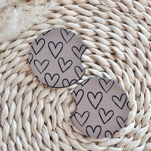 May include: Two beige car coasters with a black outline of hearts pattern.