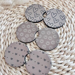 May include: Set of six brown beverage coasters with different patterns. The patterns include hearts, smiley faces, and bows.