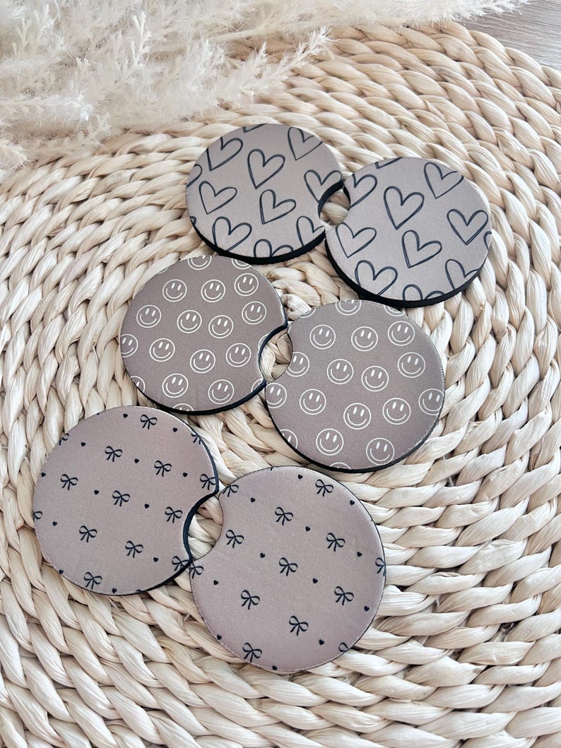 May include: Set of six brown beverage coasters with different patterns. The patterns include hearts, smiley faces, and bows.