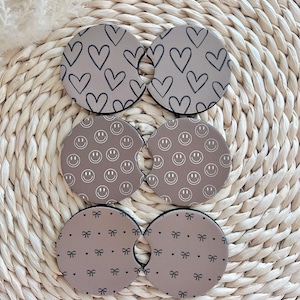 May include: Six brown round beverage coasters with different patterns. The patterns include hearts, smiley faces, and bows.