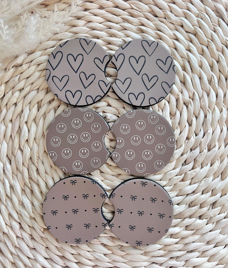 May include: Six brown round beverage coasters with different patterns. The patterns include hearts, smiley faces, and bows.