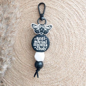 Op de afbeelding: Zwart-witte sleutelhanger met een zwarte vlinderbedel en een ronde zwarte bedel met de tekst "anti social moms club".