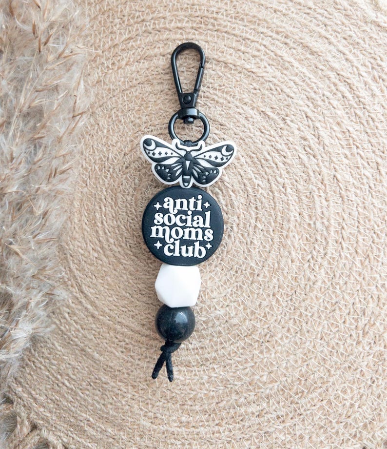Op de afbeelding: Zwart-witte sleutelhanger met een zwarte vlinderbedel en een ronde zwarte bedel met de tekst "anti social moms club".