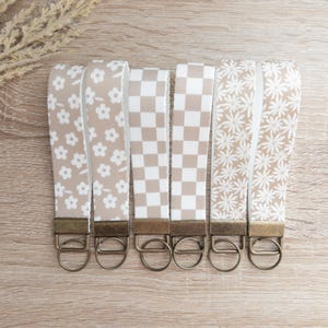 May include: Six keychains with different patterns on a light brown wooden surface. The keychains are made of beige fabric with white patterns. The patterns include small flowers, a checkered pattern, and larger flowers. The keychains have gold-colored metal rings.