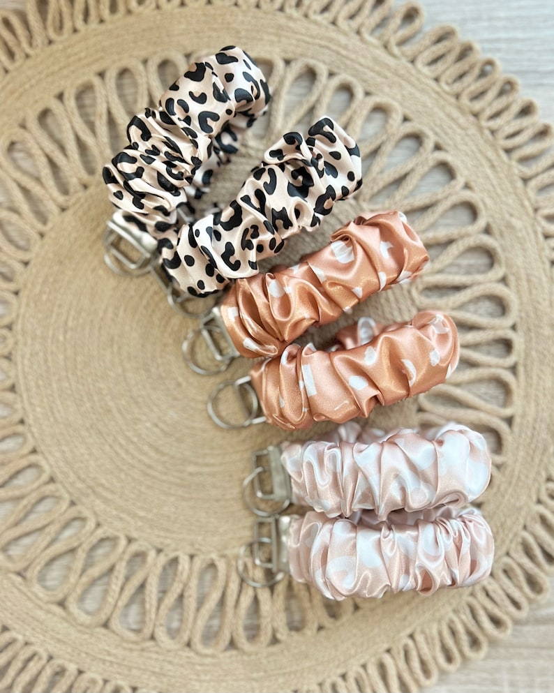 May include: Five keychains with different patterns. The keychains are made of soft, silky fabric and have a silver metal ring. The patterns include leopard print, peach with white dots, and pink with white dots.