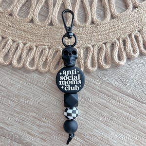 May include: Black and white silicone keychain with a skull charm, a round bead with the text "anti social moms club", a black bead, a black and white checkered bead, and a black bead.