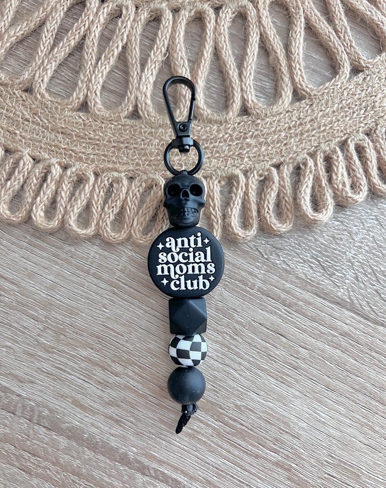 May include: Black and white silicone keychain with a skull charm, a round bead with the text "anti social moms club", a black bead, a black and white checkered bead, and a black bead.