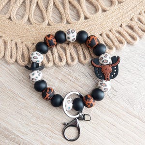 May include: A black and brown beaded keychain with a bull's head charm and a silver key ring. The beads have a variety of patterns, including leopard print and geometric designs.