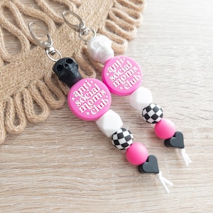 Puede incluir: Dos llaveros con forma de calavera en blanco y negro con cuentas rosas y blancas. Las cuentas tienen un patr&oacute;n de tablero de ajedrez y las cuentas rosas tienen el texto "anti social moms club".
