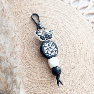 Op de afbeelding: Zwart-witte sleutelhanger met een vlinderbedel, zwarte en witte kralen en een zwarte bedel met de tekst "anti-social moms club".