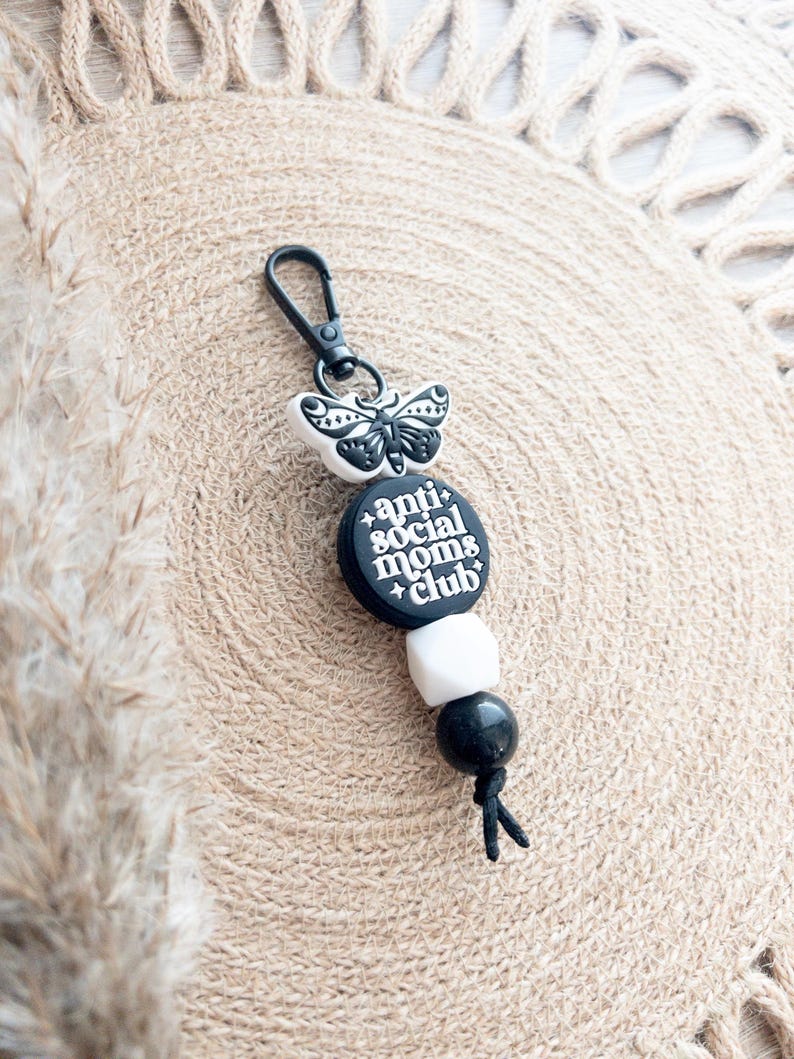 Op de afbeelding: Zwart-witte sleutelhanger met een vlinderbedel, zwarte en witte kralen en een zwarte bedel met de tekst "anti-social moms club".