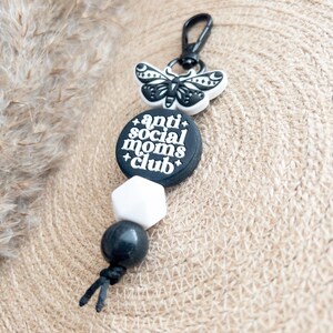Op de afbeelding: Zwart-witte sleutelhanger met een zwart-witte vlinderbedel, een zwarte kraal, een witte kraal en een zwarte bedel met de tekst "anti social moms club".