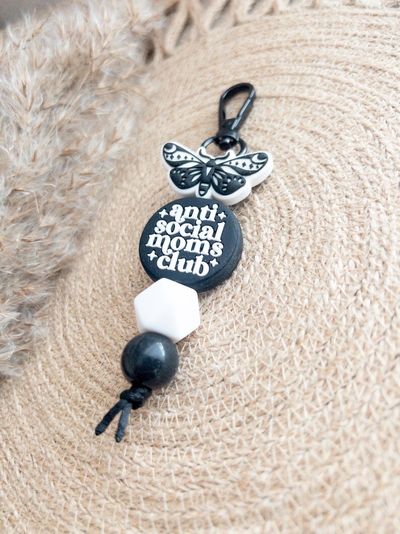 Op de afbeelding: Zwart-witte sleutelhanger met een zwart-witte vlinderbedel, een zwarte kraal, een witte kraal en een zwarte bedel met de tekst "anti social moms club".