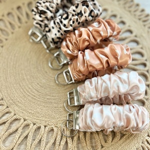 May include: A set of five keychains with different patterned fabric covers. The keychains are made of a soft, silky material and feature a silver metal ring. The fabric covers are in shades of brown, beige, and white.