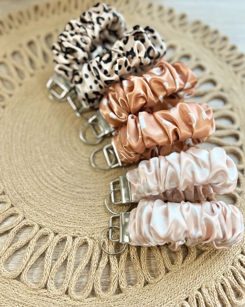 May include: A set of five keychains with different patterned fabric covers. The keychains are made of a soft, silky material and feature a silver metal ring. The fabric covers are in shades of brown, beige, and white.
