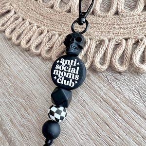 May include: Black and white silicone keychain with a skull charm and the text "anti social moms club". The keychain has a black and white checkered bead and a black bead.