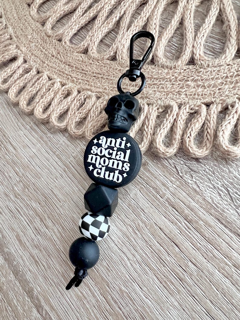 May include: Black and white silicone keychain with a skull charm and the text "anti social moms club". The keychain has a black and white checkered bead and a black bead.
