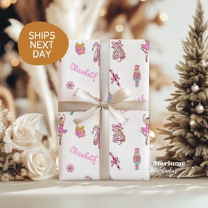 May include: Wrapped gift with a white background and a pattern of pink ballet dancers, nutcrackers, and Christmas trees. The gift is tied with a beige ribbon and bow. The name "Elizabeth" is written on the wrapping paper.