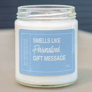 May include: A glass jar candle with a white wax interior. A light blue label reads "Smells Like Personalized Gift Message." The label also indicates the candle is 9oz and made with 100% natural soy wax, with a 50-60 hour burn time.