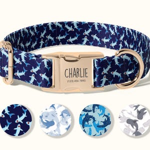 May include: A navy blue dog collar with a repeating pattern of white and light blue sharks. The gold-colored buckle is engraved with the name "Charlie" and a phone number. Four circular fabric samples with shark patterns are below the collar.