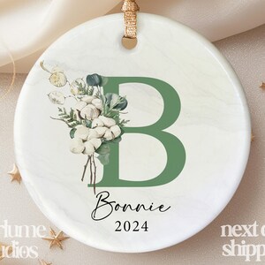 May include: White ceramic ornament with a green letter B, green leaves, and white cotton flowers. The ornament also has the name Bonnie and the year 2024.