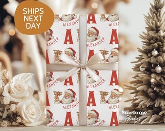 May include: Wrapped gift with a festive Santa Claus pattern in red and white. The wrapping paper features Santa holding a toy airplane, the letter "A", and the name "Alexander". A cream-colored ribbon and bow secure the gift.