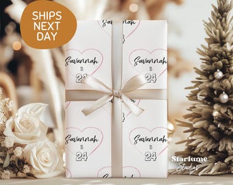 May include: A wrapped gift with a white background and a pink heart pattern with the name "Savannah" and the number "24". The gift is tied with a beige ribbon. The text "Ships Next Day" is in a gold oval.