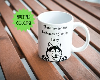 May include: White ceramic mug with a black and white Siberian Husky graphic and the text "There's no snooze button on a Siberian Husky." The mug has a curved handle and is on a wooden surface. A green starburst shape says "MULTIPLE COLORS!"