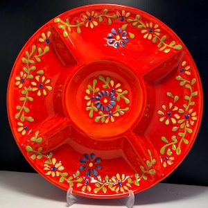Può includere: Un vivace piatto da portata in ceramica rossa con una ciotola centrale e quattro scomparti circostanti. Il piatto è decorato con un motivo floreale con fiori blu, gialli e bianchi con foglie verdi. Il piatto è rotondo.