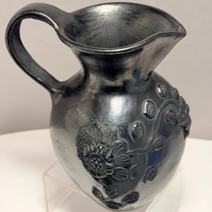 May include: A silver ceramic jug with a handle and a pouring spout. The jug has embossed floral and leaf patterns on its body. The handle is curved, and the spout is wide. The jug is displayed on a clear acrylic stand.