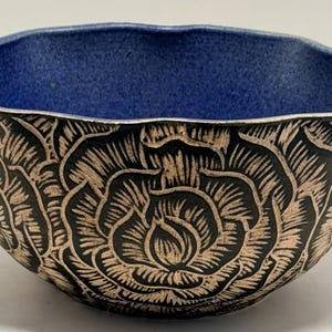 Op de afbeelding: Een keramische kom met een blauw interieur en een zwart en bruin bloemmotief aan de buitenkant. De kom heeft een geschulpte rand en een gedetailleerd gesneden rozenpatroon. De kom is waarschijnlijk handgemaakt en geschikt voor decoratief of functioneel gebruik.