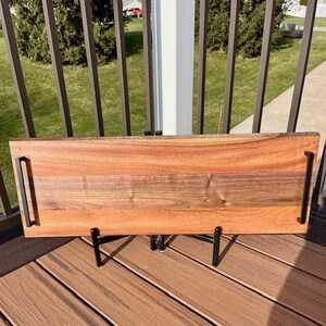 May include: A long, rectangular wooden serving tray with black metal handles. The tray is made of walnut wood and has a natural wood finish. The tray is sitting on a black metal stand.
