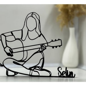 Może przedstawiać: Czarna metalowa rzeźba z drutu przedstawiająca osobę grającą na gitarze. Rzeźba stoi na białym tle, a pod nią widnieje napis "Selin".
