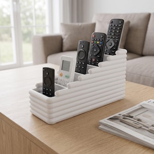 Puede incluir: Un organizador de mandos a distancia blanco de varios niveles con un diseño de barras horizontales acanaladas. El soporte rectangular cuenta con cuatro compartimentos escalonados de distintas alturas con cuatro mandos electrónicos.