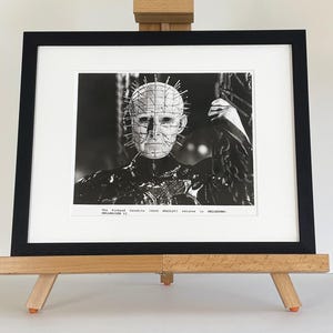 May include: Black and white framed print of a character from the horror film Hellraiser. The image features a close-up of the character's face, with pins inserted into the head. The print is displayed on a wooden easel.
