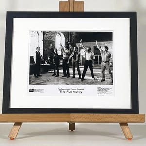 May include: A framed black and white photograph from the film "The Full Monty". The image is displayed on a wooden easel. The frame is black, and the photograph shows a group of men in a room with their arms raised in the air.