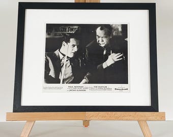 Può includere: Stampa incorniciata in bianco e nero di una scena di "The Hustler" con Paul Newman. La stampa è esposta su un piccolo cavalletto di legno con piedini arancioni. Il titolo e i crediti del film sono visibili.
