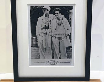 Op de afbeelding: Zwart ingelijste afdruk van een zwart-witfoto van twee mannen in lichte pakken en zonnebrillen. De afdruk wordt gepresenteerd op een kleine houten ezel. De foto komt uit de film "Another Country".