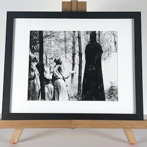 May include: Black and white framed print depicting a forest scene with figures in robes. The artwork is presented on a wooden easel. The print has a dark, mysterious atmosphere with tall trees and figures.