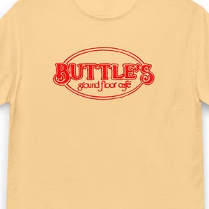 May include: A light yellow t-shirt with the red text "BUTTLE'S ground floor café" in an oval design. The shirt is hanging on a black hanger.