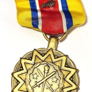 Może przedstawiać: Złoty medal wojskowy z motywem rozbłysku gwiazdy i wstążką. Wstążka jest żółta, czerwona, biała i niebieska. Medal przedstawia skrzyżowane miecze, pochodnię i gwiazdy.