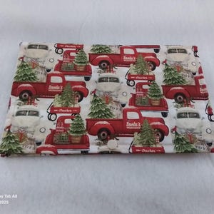 May include: A rectangular fabric item with a repeating pattern of red trucks carrying Christmas trees. The trucks have the words "Santa's Workshop" and "Dasher" printed on them. White vintage cars are also in the pattern. The background is white.