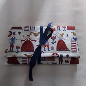 May include: A fabric roll-up case with a patriotic print featuring figures in colonial attire, stars, and the American flag. The case is tied with a navy blue ribbon.