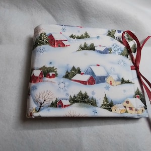 May include: A white fabric gift bag with a winter village print. The bag has a red ribbon tied around it.
