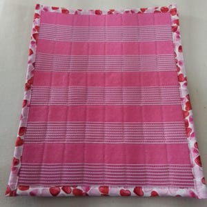 Op de afbeelding: Een roze gewatteerde placemat met horizontale witte strepen. De rand heeft een patroon van rode harten op een witte achtergrond. De placemat is rechthoekig en lijkt handgemaakt.