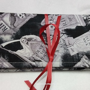 May include: A black and white fabric pouch with a spooky graveyard print. The pouch is tied with a red ribbon and features the text "RIP", "Dust to Dust", and "I'll Be Back".