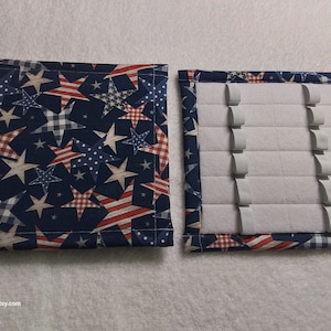 May include: A two-piece jewelry organizer with a patriotic theme. The exterior features a navy blue fabric with white, red, and blue stars. The interior has a padded gray surface with elastic bands for securing jewelry.
