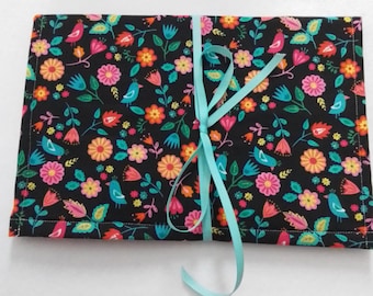 Pode incluir: Um organizador de tecido retangular com fundo preto e um padrão colorido de flores rosas, laranjas e turquesas, além de pequenos pássaros. O item está dobrado e preso no centro com uma fita azul-clara amarrada em um laço simples.